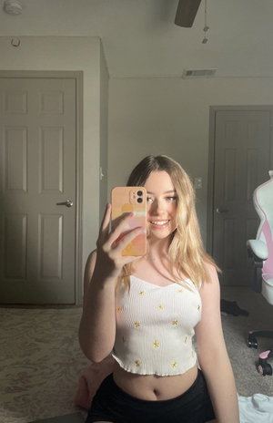 Blonde woman in white crop top taking mirror selfie in bedroom.