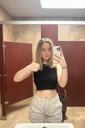 Woman in black crop top and beige shorts taking mirror selfie in bathroom.