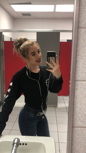 Woman with blonde hair in bun, wearing black long-sleeve top and jeans, taking mirror selfie in