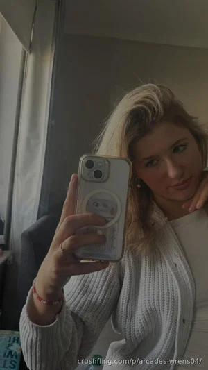 Woman with blonde hair in white knit cardigan taking mirror selfie indoors.