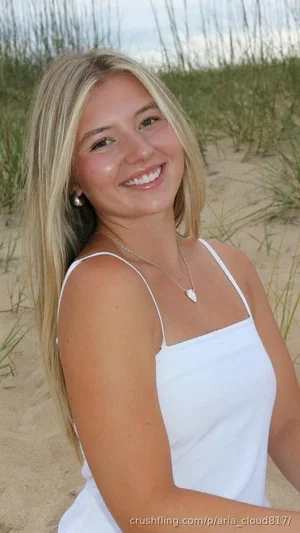 Blonde woman in white top smiling on sandy beach with grass behind.
