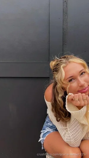 Woman with blonde hair in off-shoulder sweater and denim shorts, posing against dark wall.