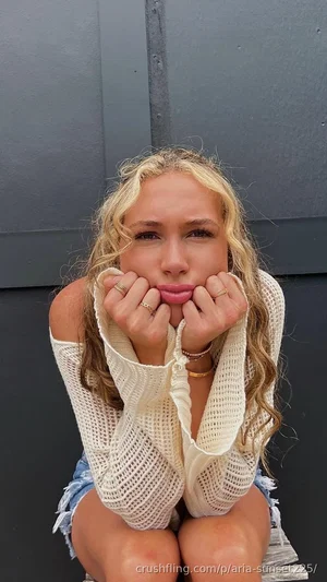 Blonde woman in off-shoulder knit top and denim shorts, sitting against gray wall.