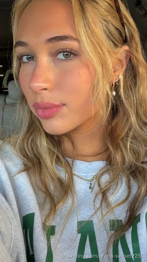 Woman with blonde wavy hair wearing a gray sweatshirt with green text, sitting in a car.