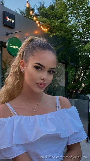 Woman with blonde ponytail in white off-shoulder top, outdoors near restaurant with string lights.