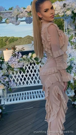 Woman in beige ruffled dress standing on deck with floral arch, outdoor setting.