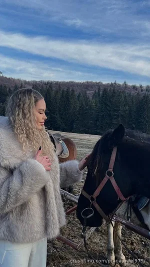 Woman with curly blonde hair in a fur coat petting a horse outdoors.
