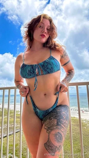 Woman in blue patterned bikini with tattoos, standing on balcony overlooking beach.