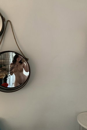 Dark-haired woman taking topless mirror selfie indoors.