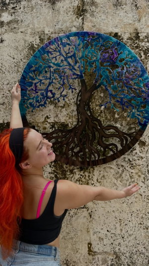 Woman with red hair in black top and jeans poses near colorful tree mural on stone wall.