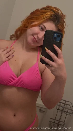 Woman with red hair in pink bikini taking mirror selfie in bathroom.