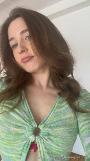 Woman with brown hair in a green patterned top, showing a red bra strap, indoors.