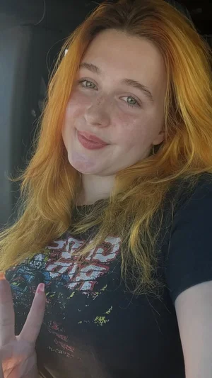 Woman with long red hair wearing dark graphic t-shirt, smiling in close-up selfie.