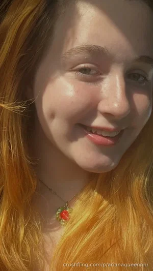 Woman with long red hair and a floral pendant necklace, smiling in sunlight.