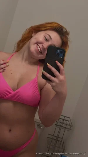 Woman with red hair in pink bikini taking mirror selfie in bathroom.