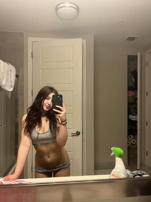 Woman in gray sports bra and shorts taking mirror selfie in bathroom.