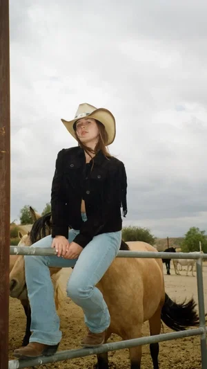 Woman in cowboy hat and jeans sitting on a horse in a rural outdoor setting.