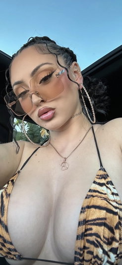 Woman in tiger-print bikini top wearing large hoop earrings and sunglasses, taking selfie in car.