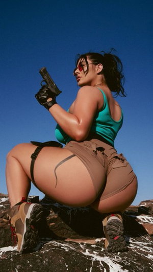 Woman in teal top and brown shorts posing with gun on rocky terrain under blue sky.