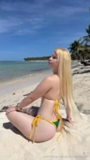 Blonde woman in yellow bikini sitting on sandy beach, facing side, tropical background.