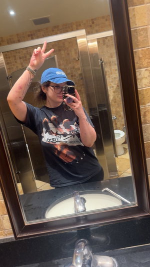Woman in blue cap and glasses taking mirror selfie in bathroom.