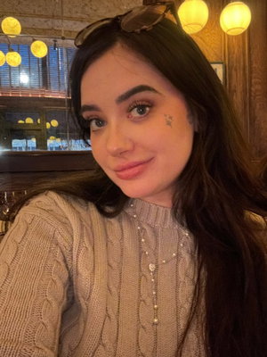 Woman with long dark hair in beige sweater taking selfie indoors.