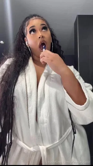 Woman with long curly hair in white robe brushing teeth indoors.
