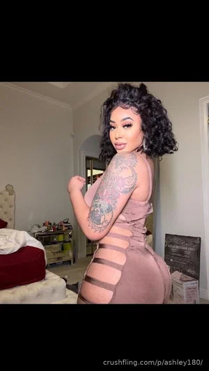 Woman with curly hair and arm tattoo wearing a cut-out dress in a bedroom.