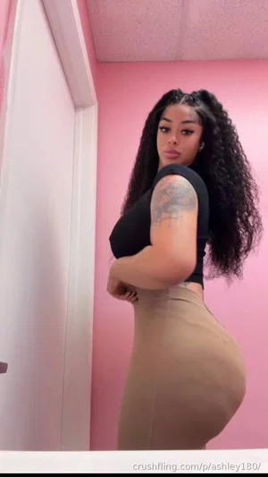 Woman with curly hair in black top and beige skirt posing in pink room.