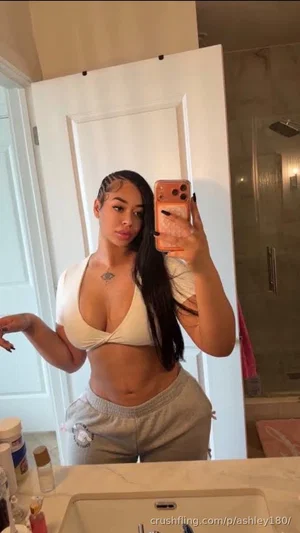 Woman in white crop top and gray sweatpants takes mirror selfie in bathroom.