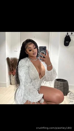 Woman in white fringed top kneeling in bedroom, holding phone for mirror selfie.