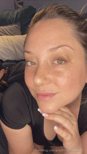 Woman with freckles and green eyes in black top, resting on bed, hand under chin.
