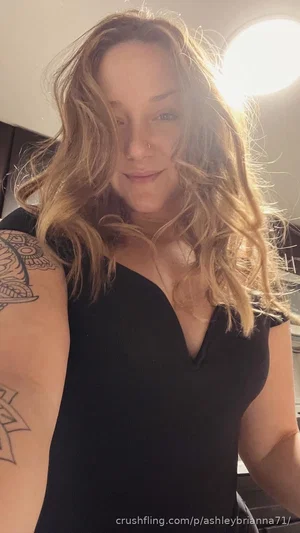 Woman with wavy blonde hair, tattooed arm, wearing black top, taking mirror selfie indoors.