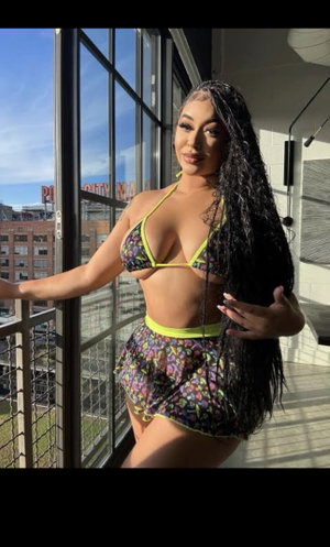 Woman in floral bikini with neon trim poses on balcony, long dark hair, city view behind.