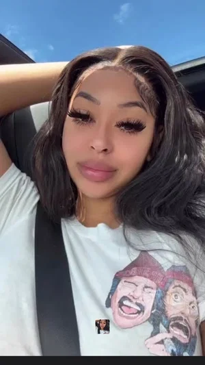 Woman with long dark hair wearing white graphic t-shirt, sitting in car, hand on head.