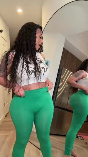 Woman with curly hair in white crop top and green leggings, standing in front of mirror indoors.