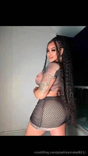 Woman with long braids in sheer top and mesh skirt, posing indoors.