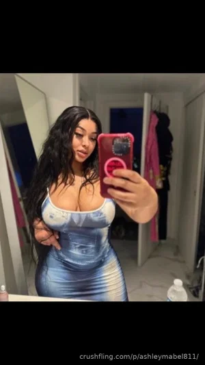 Woman with long dark hair in a tight blue dress taking mirror selfie in a room.