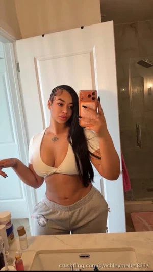Woman in white top and gray sweatpants taking mirror selfie in bathroom.