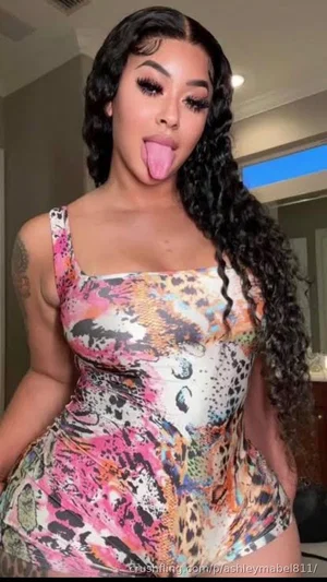 Woman with long curly hair in colorful patterned bodysuit, posing indoors with tongue out.