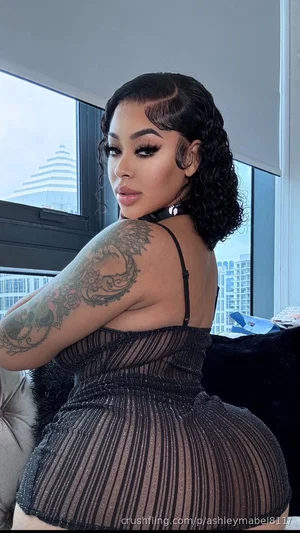 Woman in black sheer dress with tattoos, posing indoors near window.