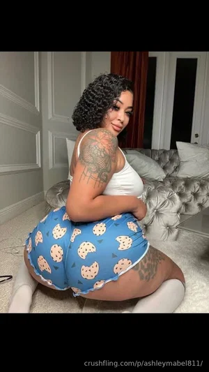 Woman with curly hair and tattoos wears white top and blue patterned shorts, posing on carpet in