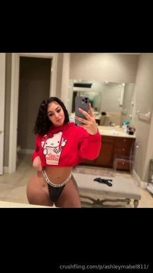 Woman in red crop hoodie and black thong takes mirror selfie in bathroom.