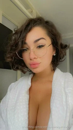 Woman with curly brown hair and glasses wearing an open white robe indoors.