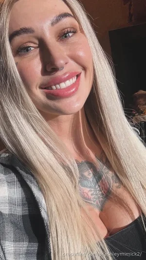 Woman with long blonde hair, plaid shirt, chest tattoo, smiling indoors.