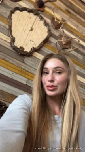 Woman with long blonde hair in a white top, indoors with wooden wall decor.
