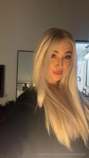Woman with long blonde hair in black top, indoors, mirror visible behind.