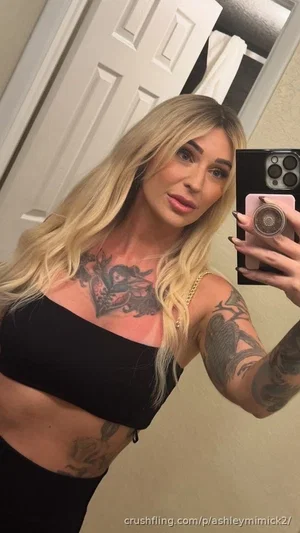 Blonde woman with tattoos in black crop top and pants, taking mirror selfie indoors.