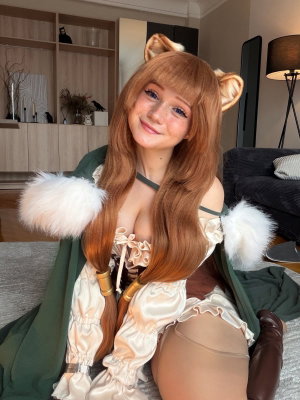 Woman in cosplay outfit with long brown hair, animal ears, and green cloak sitting indoors.