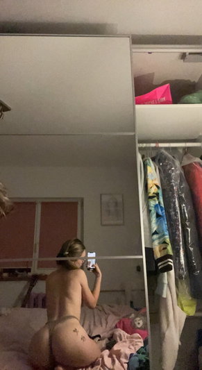 Woman with glasses taking mirror selfie in bedroom, wearing thong, bare back visible.